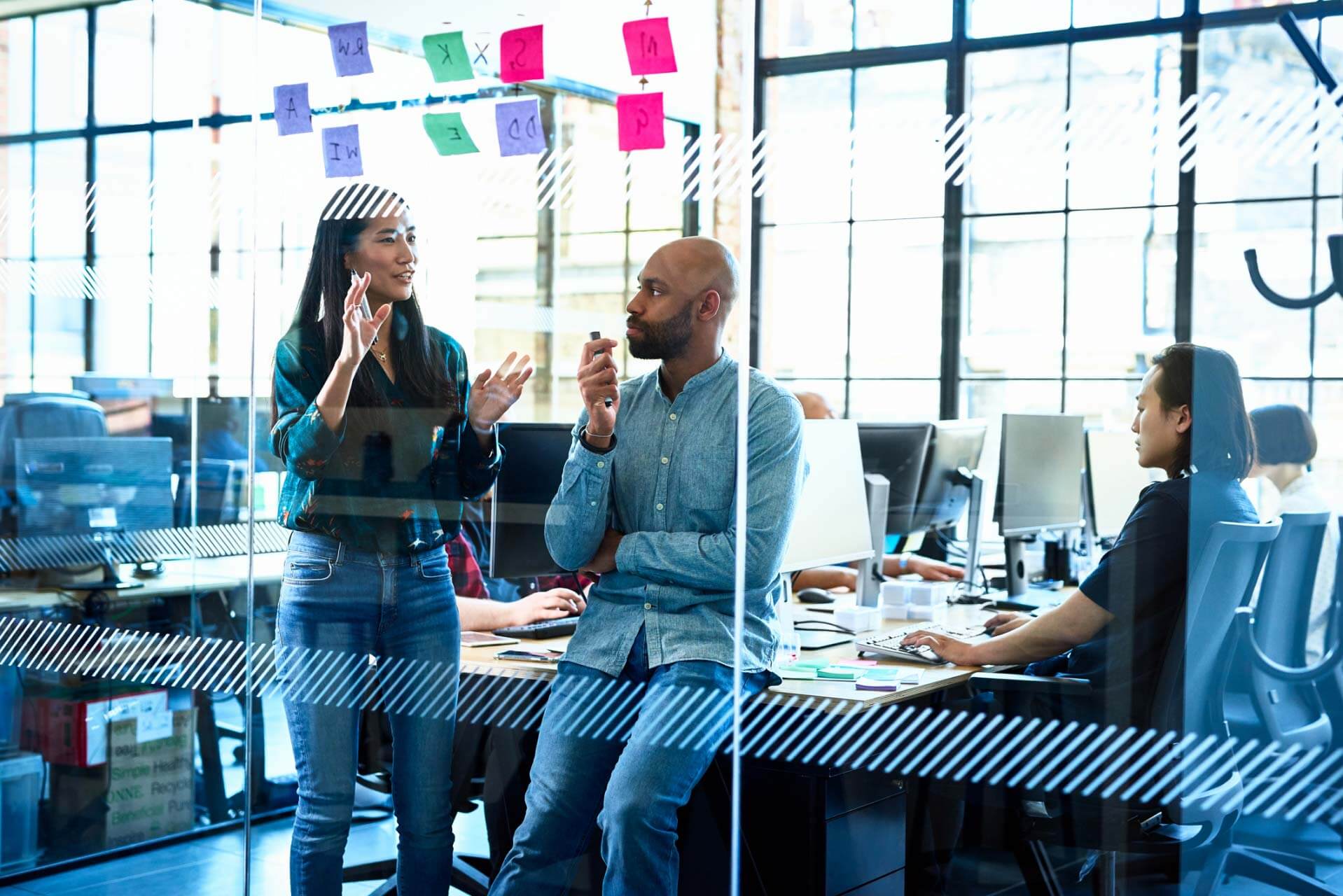 Two coworkers standing by desks and talking in an office setting.